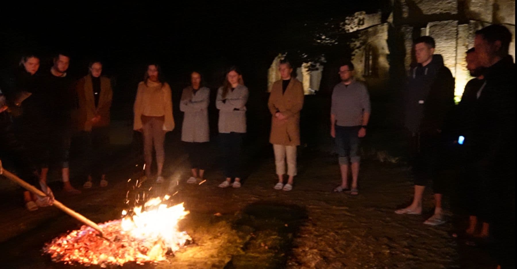 A group standing in a circle at night as the fire is raked into a bed of glowing embers