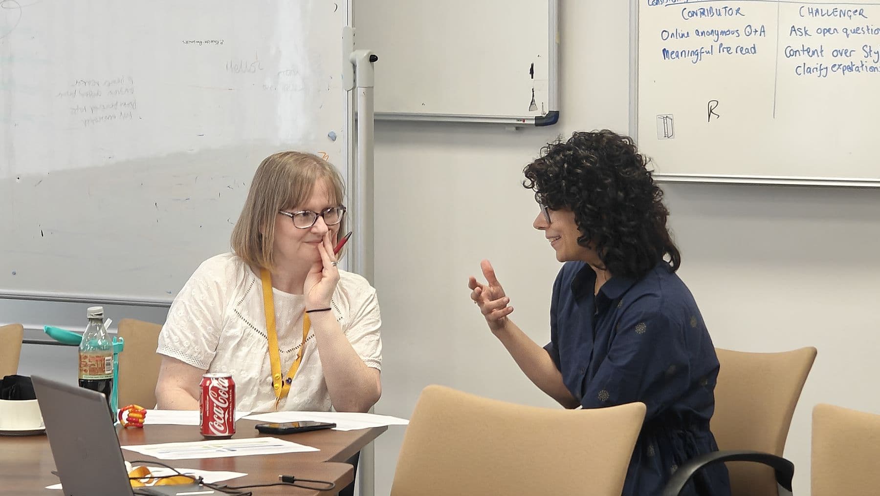 Two women in a coaching conversation at a table, one listening closely as the other talks