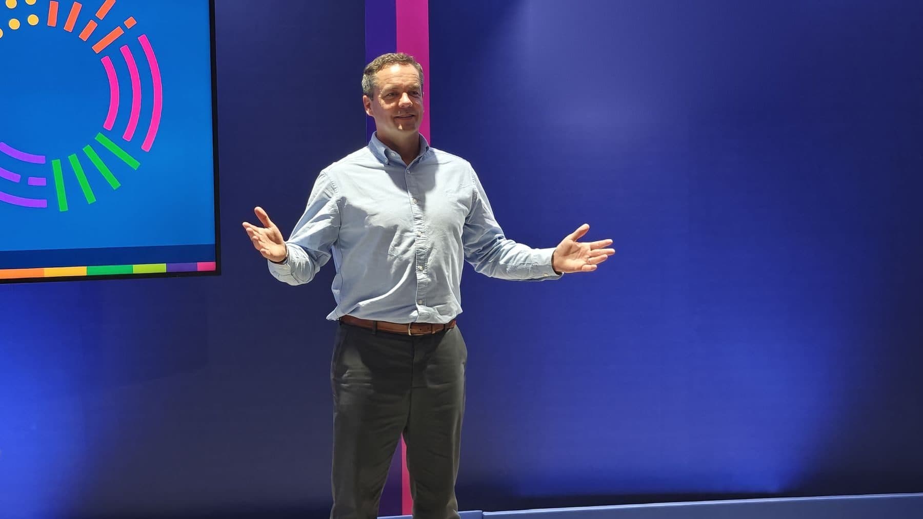 A speaker on stage in front of a large screen, arms open to the audience