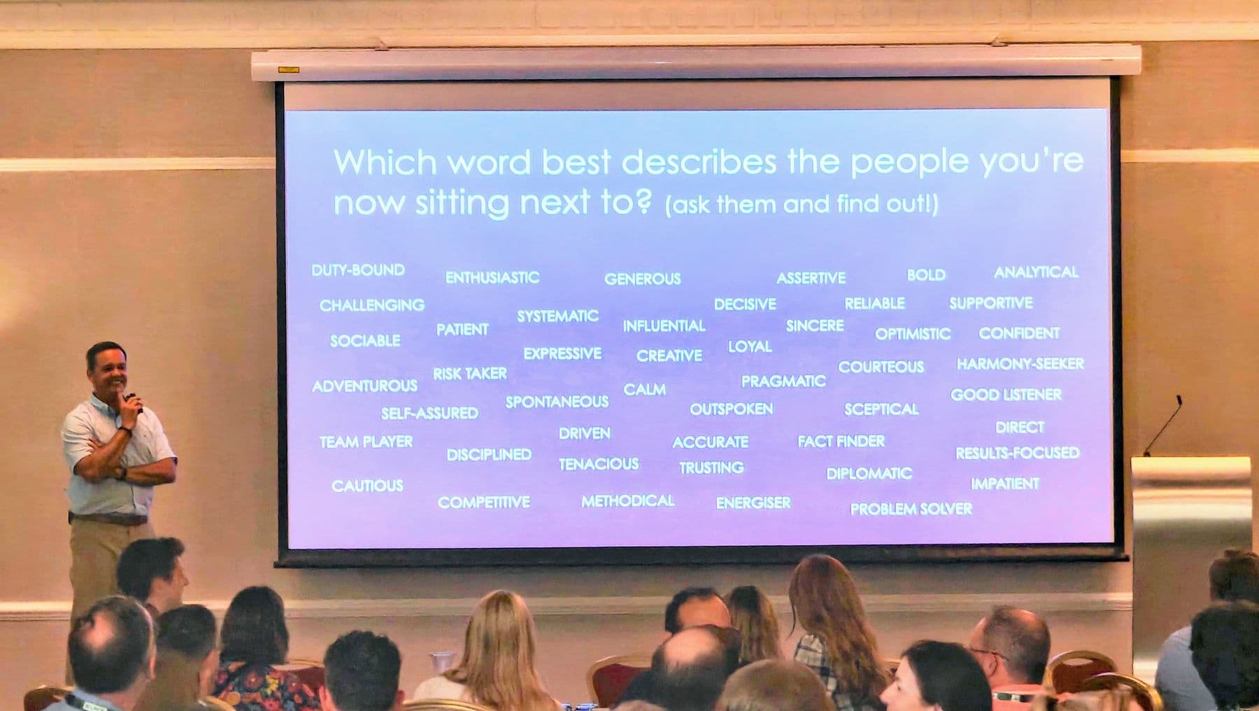 A speaker presenting to a conference audience in front of a slide of personality words